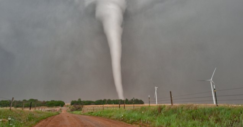A tornado descends from dark storm clouds near a dirt road, with wind turbines visible in the foreground.
