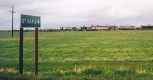 A green field with a sign pointing to "St Marie" and a distant view of a small town under a cloudy sky.