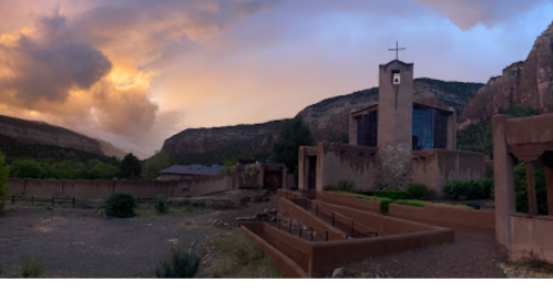 A panoramic view of a church surrounded by mountains and dramatic clouds at sunset.