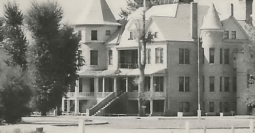 Historic building with multiple towers, large windows, and a porch, surrounded by trees and a grassy area.