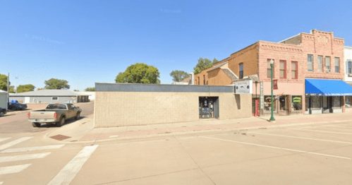 Street view of a small town with a brick building, a blue awning, and a parked truck on a sunny day.