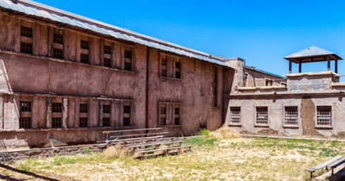 Abandoned prison building with barred windows and a watchtower under a clear blue sky. Overgrown grass in the courtyard.