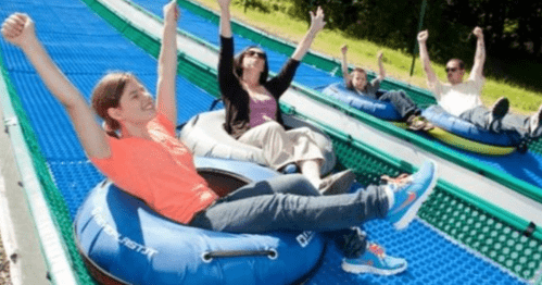 Four people on inflatable tubes sliding down a blue, outdoor slope, all with arms raised in excitement.