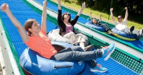 Four people on inflatable tubes sliding down a blue, outdoor slope, all with arms raised in excitement.