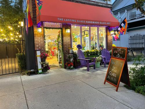 A colorful storefront with a sign reading "The Laffing Glass Handmade Glass Art," featuring outdoor seating and decorations.