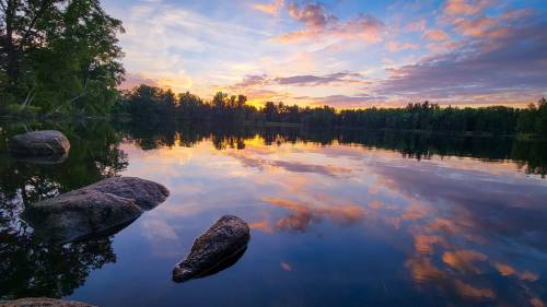 A serene lake at sunset, with colorful clouds reflecting on the water and rocks in the foreground.