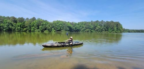 A person paddles a kayak on a calm lake surrounded by lush green trees under a clear blue sky.