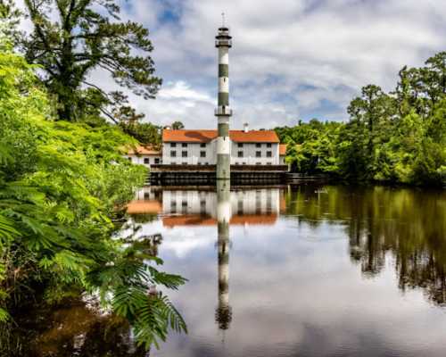 A tall lighthouse reflects in calm waters, surrounded by lush greenery and a historic building in the background.