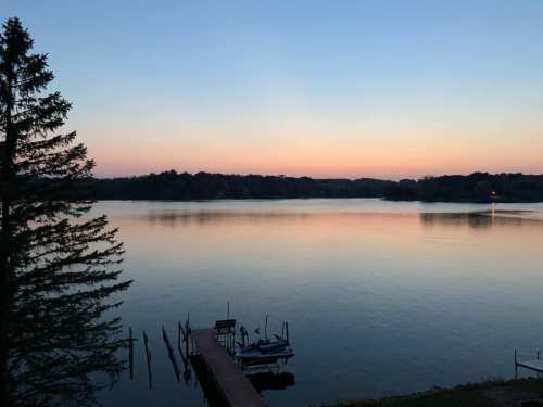 A serene lake at dusk, with a calm surface reflecting the pastel sky and a small dock in the foreground.