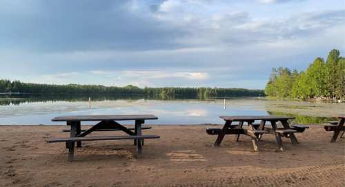 A serene lakeside view with picnic tables on sandy shore, surrounded by trees and calm water under a cloudy sky.
