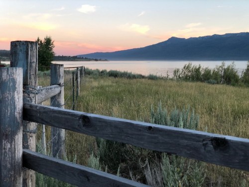 A serene lakeside view at sunset, featuring a wooden fence and lush greenery with mountains in the background.