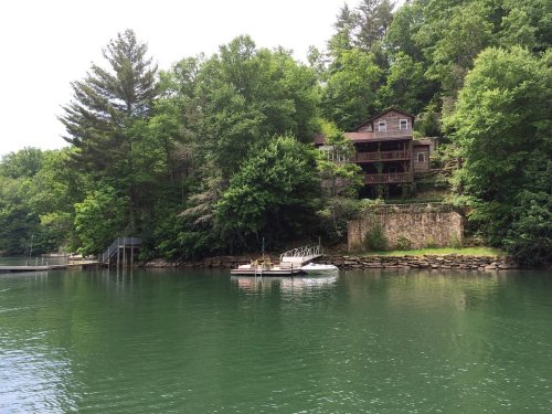 A serene lake scene featuring a house on a hillside surrounded by lush greenery and a small boat docked nearby.