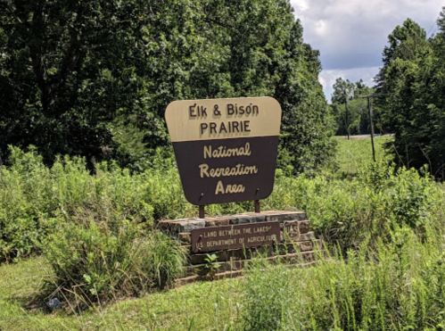 Sign for Elk & Bison Prairie National Recreation Area, surrounded by greenery and trees under a partly cloudy sky.