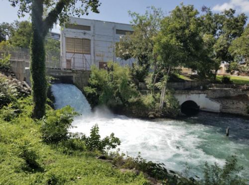 A flowing river with a small waterfall beside a building, surrounded by lush greenery and trees.
