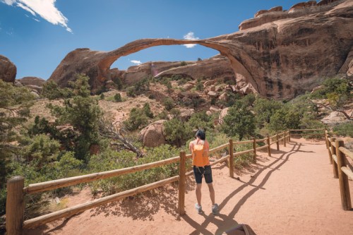 A person stands on a path, taking a photo of a large rock arch surrounded by desert landscape and greenery.