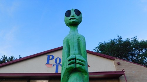 A tall green alien statue stands in front of a building with the word "Pot" on it, under a clear blue sky.