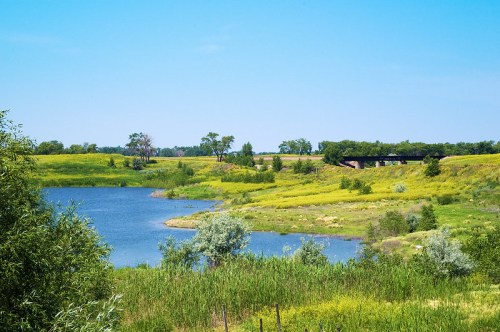 A serene landscape featuring a blue lake, lush green fields, and a distant bridge under a clear blue sky.