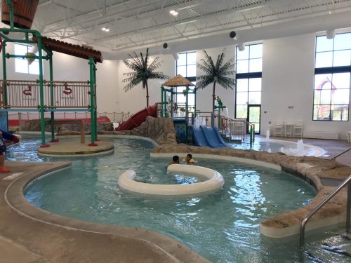 Indoor water park featuring a lazy river, slides, and palm trees, with children playing in the water.