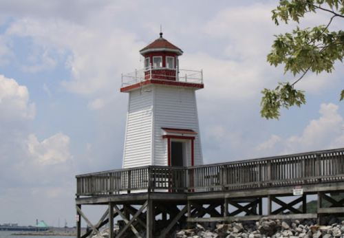A white lighthouse with a red roof stands on a wooden pier, surrounded by a cloudy sky and rocky shoreline.