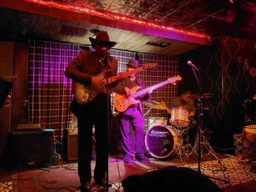 A band performs on stage with a guitarist in a hat, a bassist, and a drummer, illuminated by purple lights.