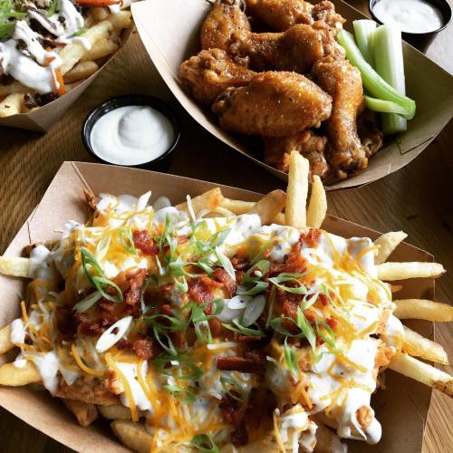 A close-up of loaded fries topped with cheese, bacon, and green onions, alongside chicken wings and celery sticks.
