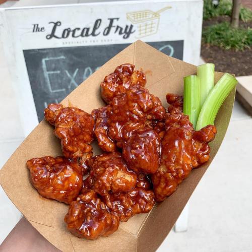 A hand holding a paper tray filled with sticky, glazed chicken wings and celery sticks, with a sign in the background.