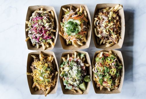 A variety of loaded fries in cardboard trays, topped with colorful ingredients like cheese, vegetables, and sauces.