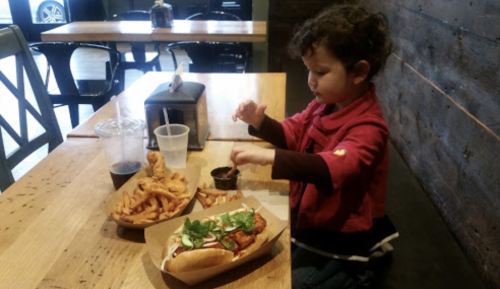 A child sits at a table, enjoying a meal with a hot dog, fries, and a drink in a casual dining setting.