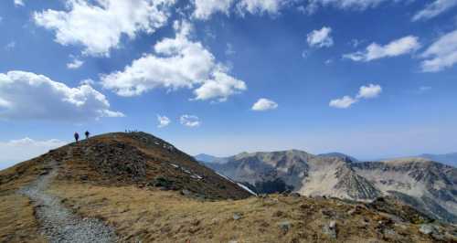 A scenic mountain ridge with hikers, under a blue sky with fluffy clouds and distant peaks in the background.