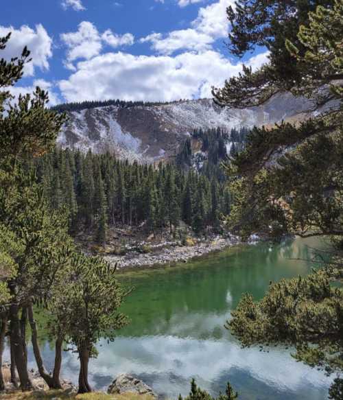 A serene lake surrounded by evergreen trees and mountains, under a partly cloudy sky.