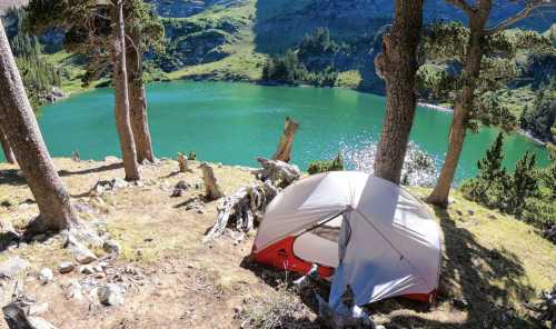 A tent set up near a vibrant green lake, surrounded by trees and mountains under a clear blue sky.