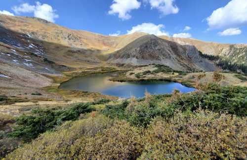 A serene mountain landscape featuring a calm lake surrounded by rocky hills and scattered greenery under a blue sky.