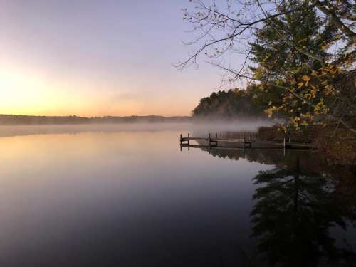 A serene lake at dawn, with mist rising and trees reflecting on the calm water.
