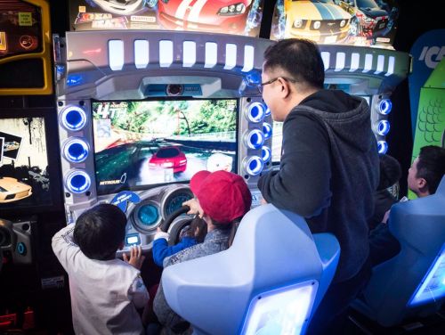 Two children and an adult play a racing arcade game, focused on the screen displaying a car racing scene.