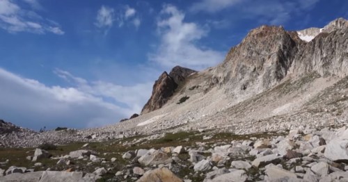 A rocky mountain landscape under a blue sky with scattered clouds and a grassy foreground.
