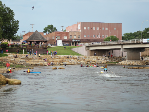 A river scene with people kayaking and paddleboarding, a bridge in the background, and a park area with onlookers.