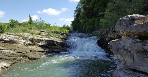 A serene waterfall cascades over rocky terrain, surrounded by lush greenery and a clear blue sky.