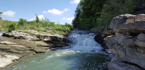 A serene waterfall cascades over rocky terrain, surrounded by lush greenery and a clear blue sky.
