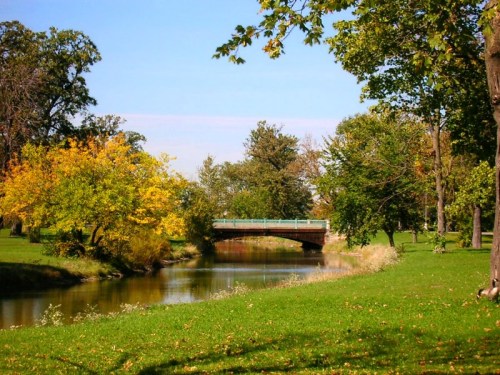 A serene park scene featuring a calm river, a small bridge, and vibrant trees in autumn colors under a clear blue sky.