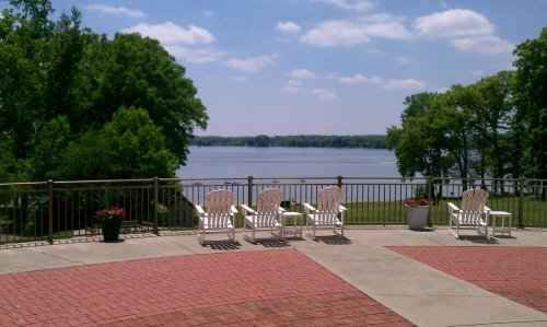 A serene lakeside view with white chairs on a patio, surrounded by greenery and a clear blue sky.