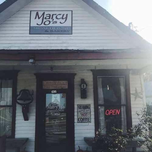 A quaint white building with a sign reading "Marcy Jo's Mealhouse & Bakery" and an "Open" neon sign in the window.
