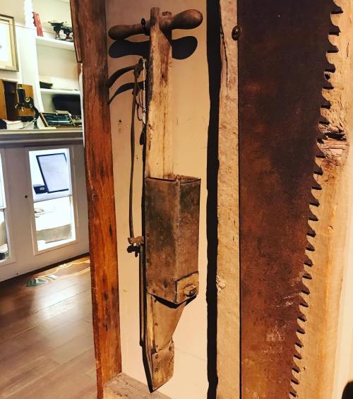 An old wooden tool with a metal box and handle, displayed on a wall alongside a saw.