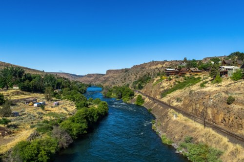 A winding river flows through a dry landscape with hills, trees, and houses along the banks under a clear blue sky.