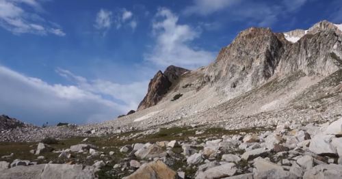 A rocky mountain landscape under a blue sky with scattered clouds and patches of green vegetation.