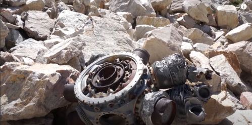 A rusted mechanical component lies among large, jagged rocks in a rugged outdoor setting.