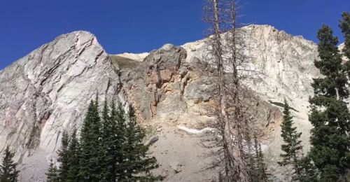 A rocky mountain landscape with tall trees in the foreground and a clear blue sky above.