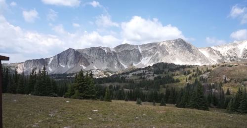 A scenic view of rugged mountains under a blue sky, surrounded by green trees and grassy fields.