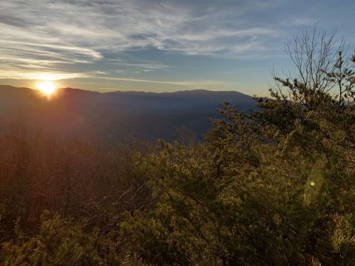 Sunset over a mountainous landscape, with trees in the foreground and a colorful sky filled with clouds.