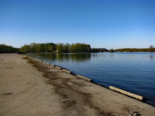 A calm lake with a sandy shore, surrounded by green trees under a clear blue sky.