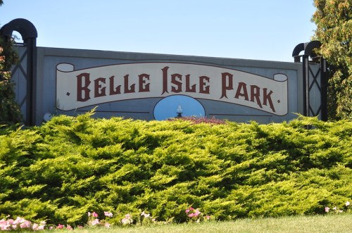 Sign for Belle Isle Park surrounded by green shrubs and flowers under a clear blue sky.